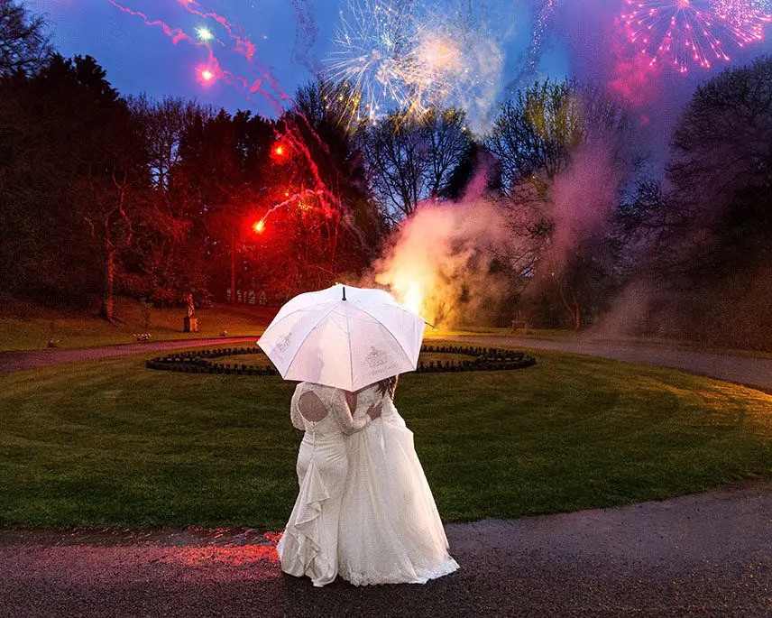 Two brides in wedding dresses sharing a moment under a white umbrella as colourful fireworks light up the evening sky in a garden.